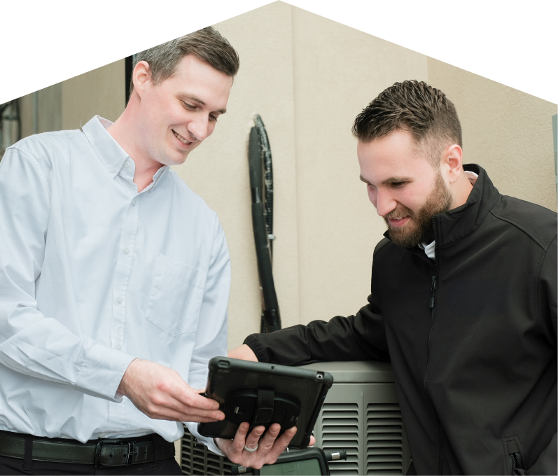 A furnaceguys technician in a white shirt shows service details on a tablet to a smiling client in a black jacket, standing beside an AC unit outside a beige building.