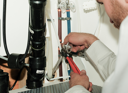 Using two pipe wrenches, a furnaceguys professional plumber services a pipe connection near a vertical flue or vent pipe.